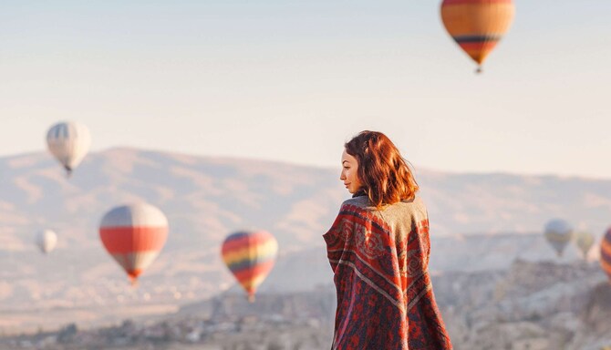 A woman wrapped in a blanket watching hot air balloons over the desert.