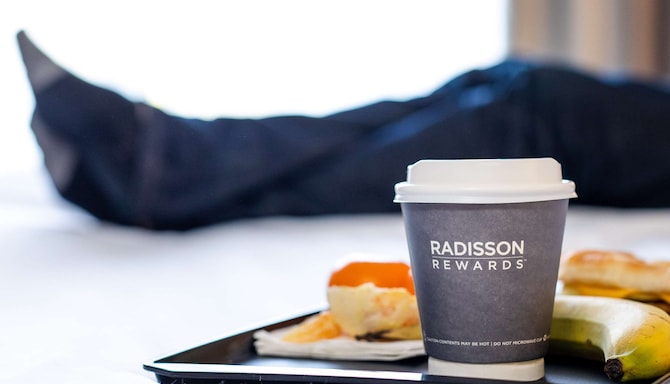 A tray with a to-go coffee cup and breakfast foods rests on a white bed