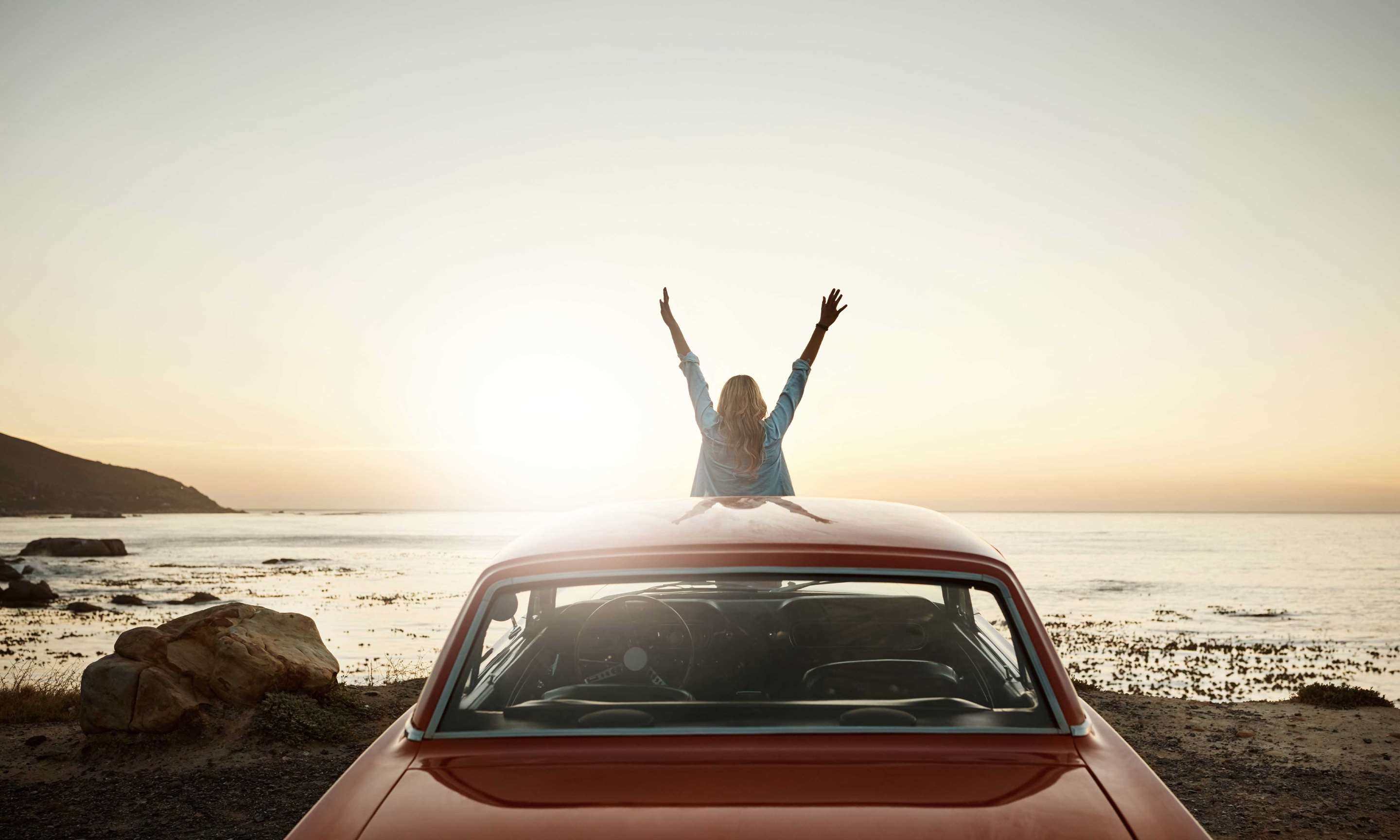 A woman with arms raised, sitting on the hood of her car while looking at the ocean sunset.