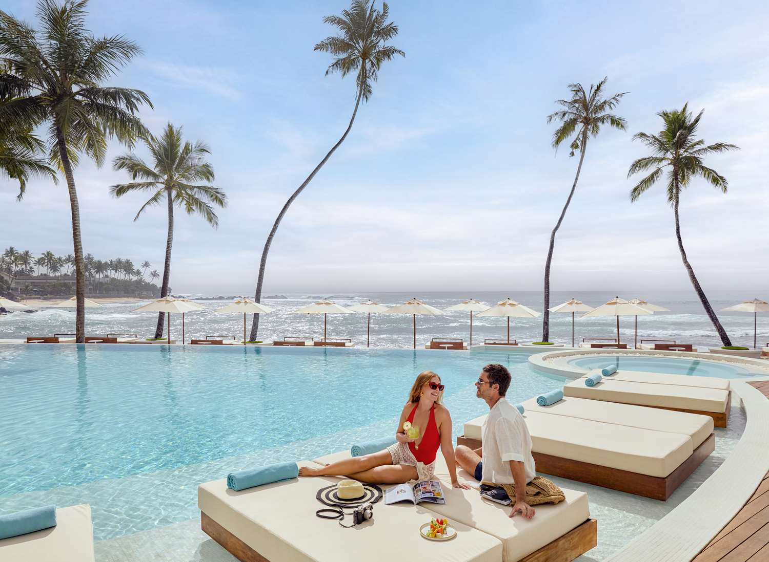 A couple on a daybed at the edge of a curving infinity pool, with white parasols, palms and the sea beyond.