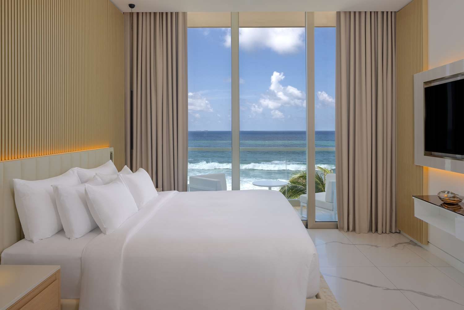 A white bedroom with a wide bed and a floor-to-ceiling window onto a balcony over the sea.