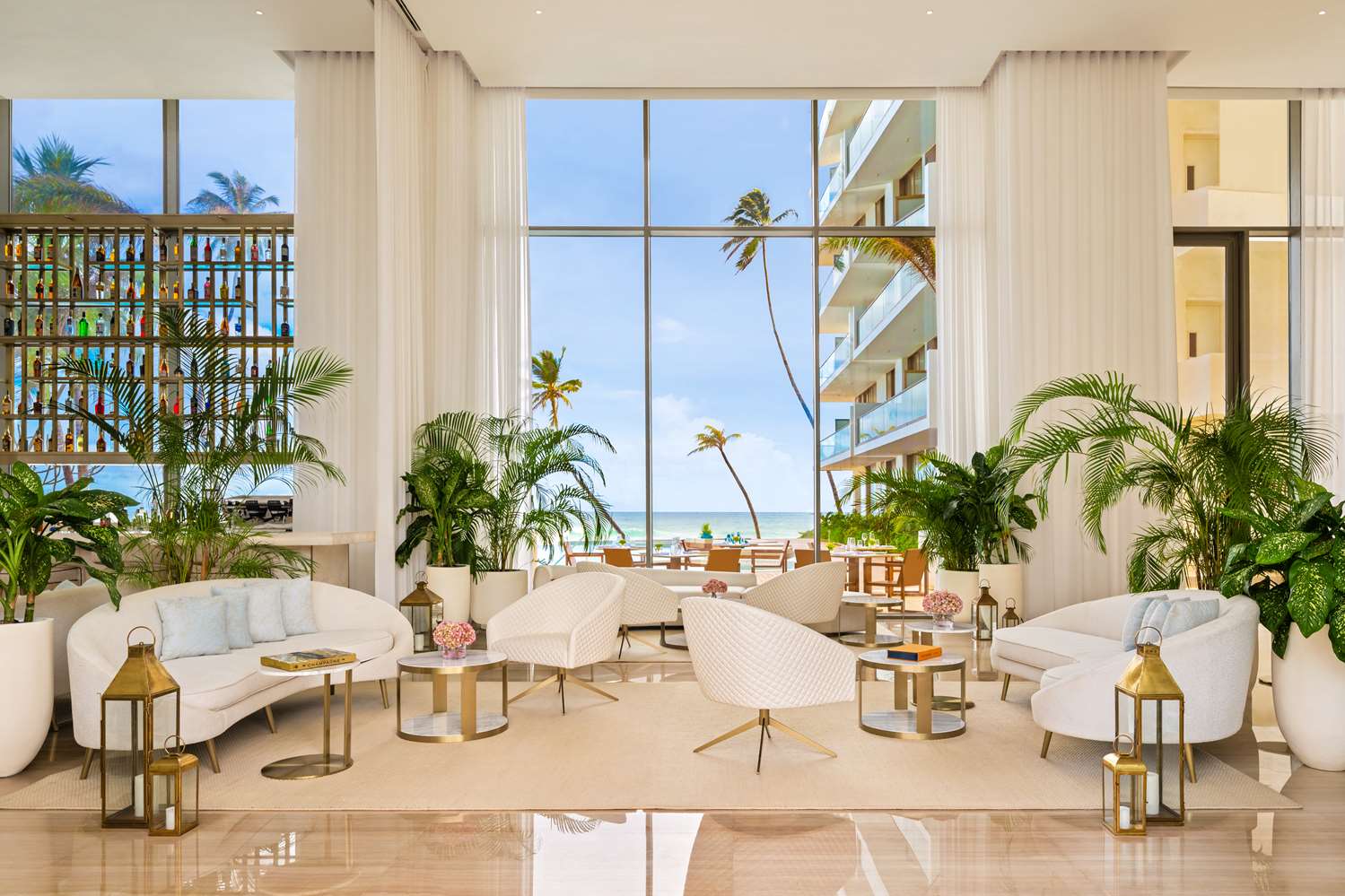 A double-height lounge with white armchairs and potted palms, its tall windows looking out to the sea.