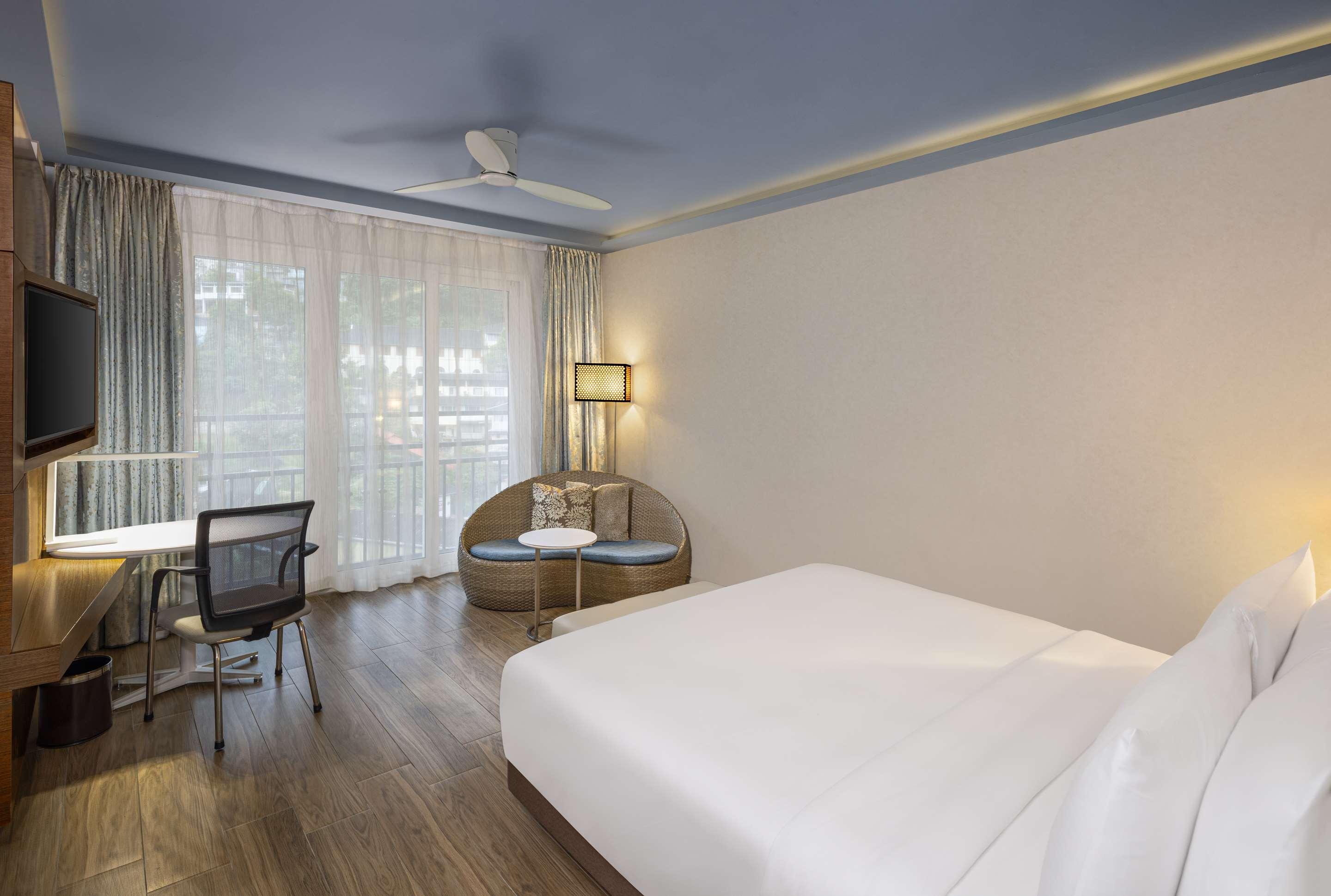 A Deluxe Room: a large white bed, a rattan armchair by a window hung with sheer curtains, a desk and a ceiling fan over a timber floor