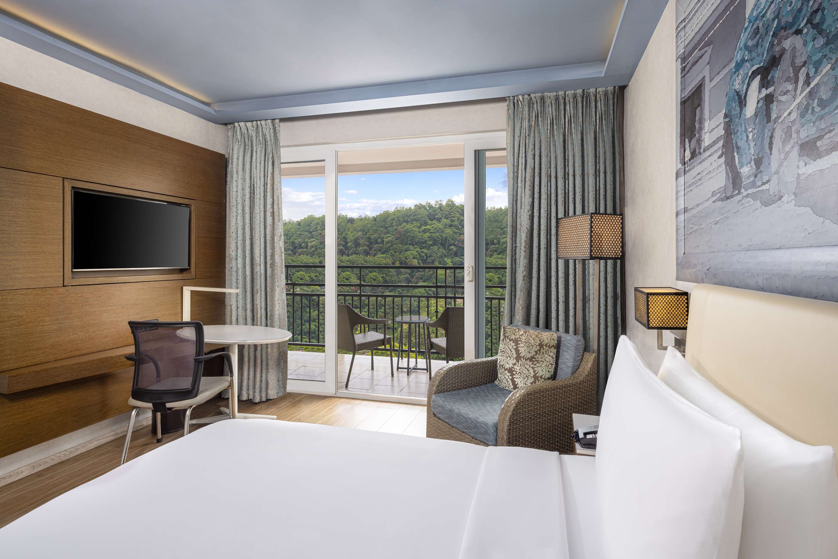 A Deluxe Room - Lake View: a white bed, a wicker armchair, a round table and a glass door open to a balcony facing wooded hills