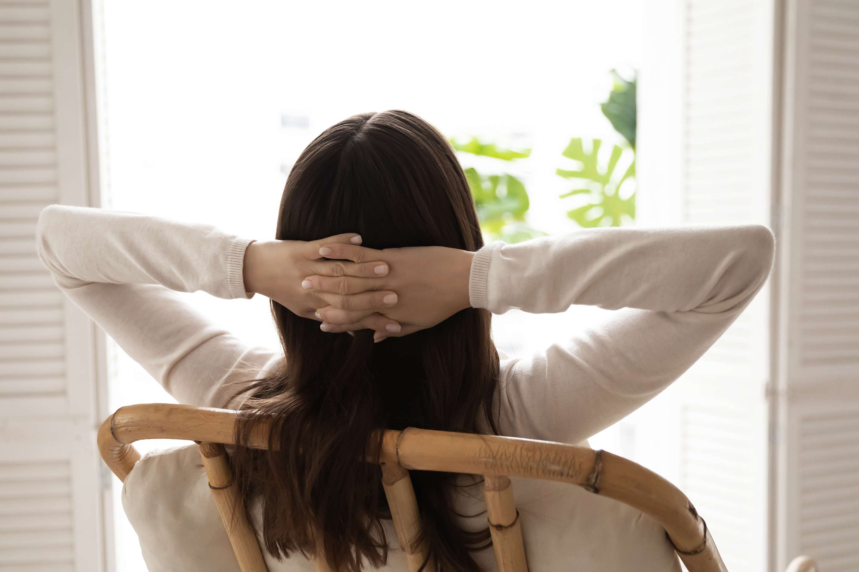 A woman sitting in a chair, looking out a window with her hands behind her head.