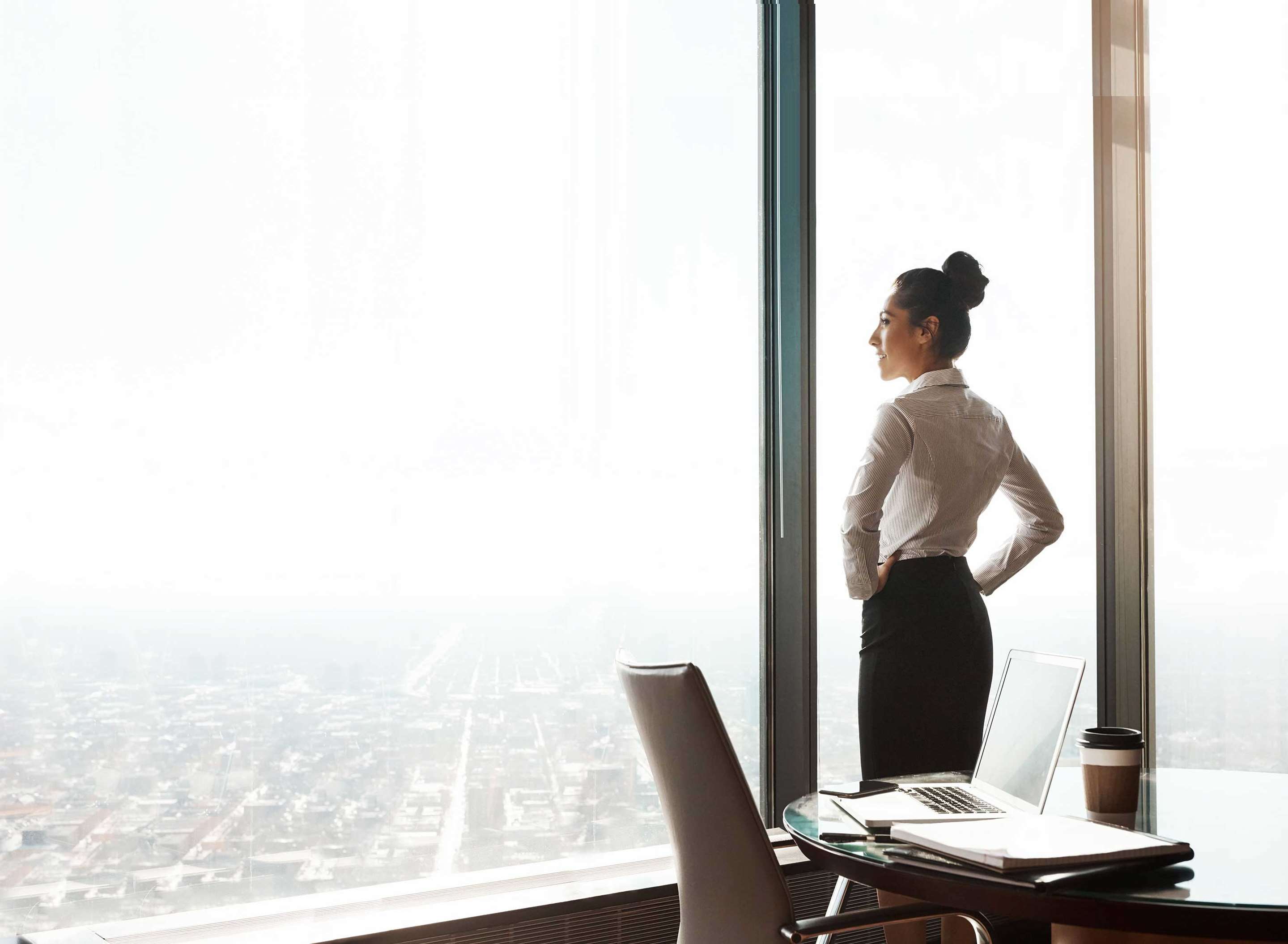 A businesswoman in an office, looking out window at city skyline.